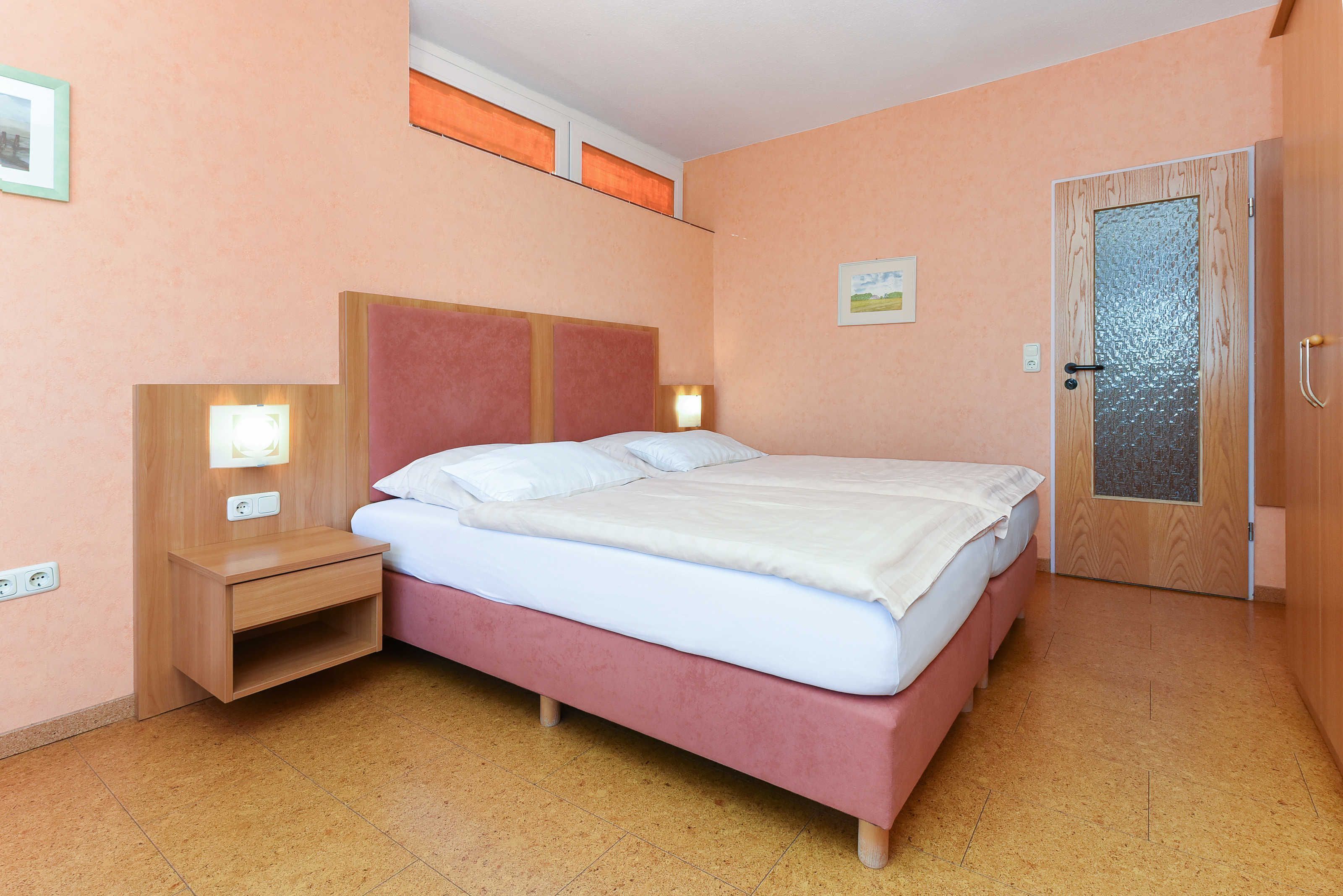 A bedroom with a bed, bedside tables, a nightstand, a door, and a wall-mounted light fixture.