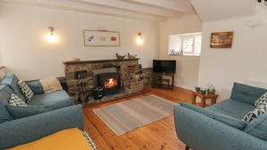 A cozy living room with a stone fireplace, two sofas, a television, and a wall-mounted shelf. - Little Barn Cottage (Newport)
