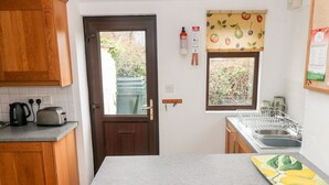 Una cocina con puerta de madera, ventana con diseño de frutas, un tostador, una tetera y un escurreplatos.