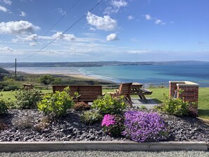 A patio with wooden benches and a brick structure overlooking a beach and ocean. - Ysgubor Ddegwm Bach (Beaumaris)