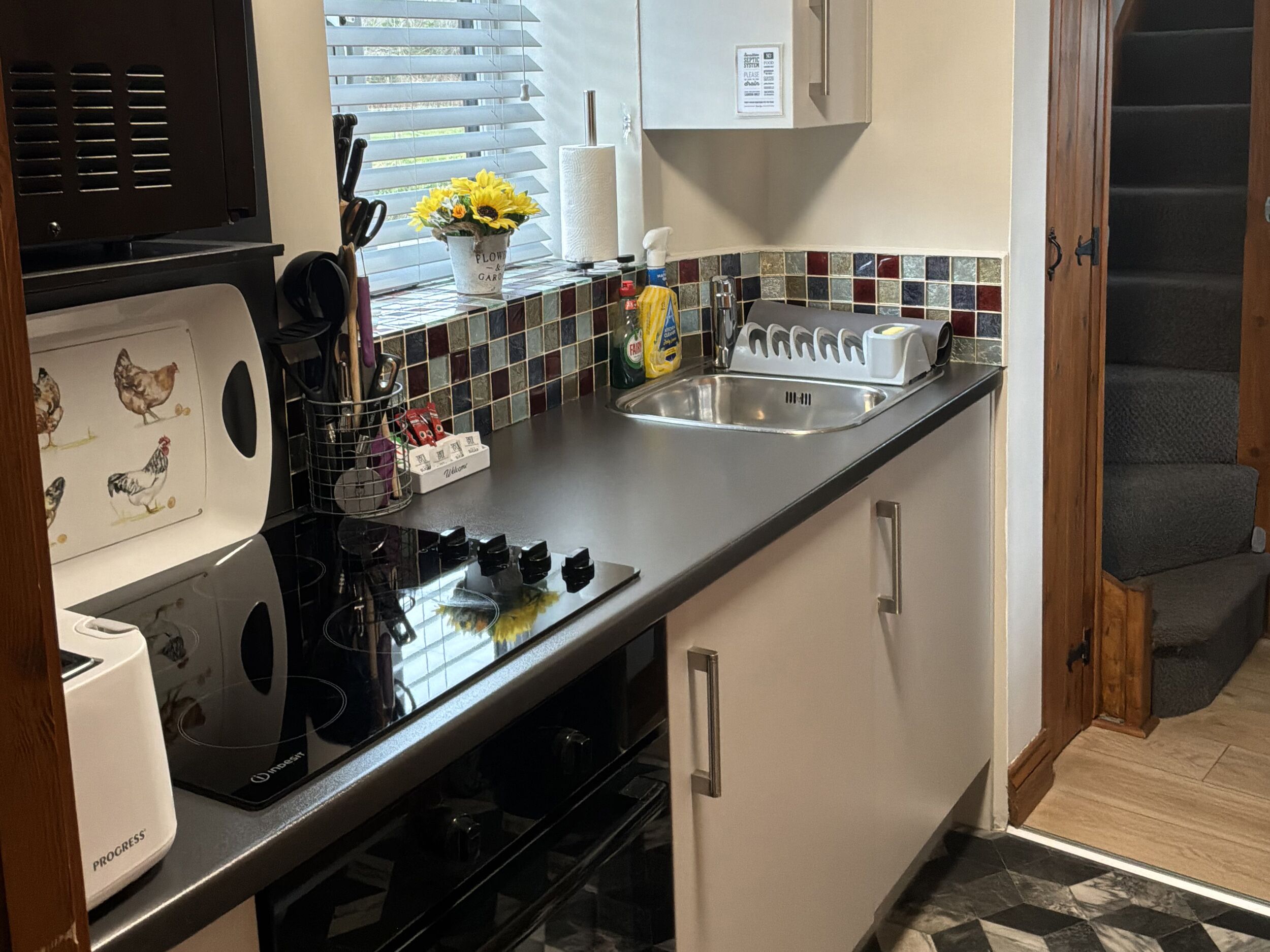 A compact kitchen with a stove, sink, and a small countertop area. There is a window with blinds, a vase with flowers, and a tiled backsplash.