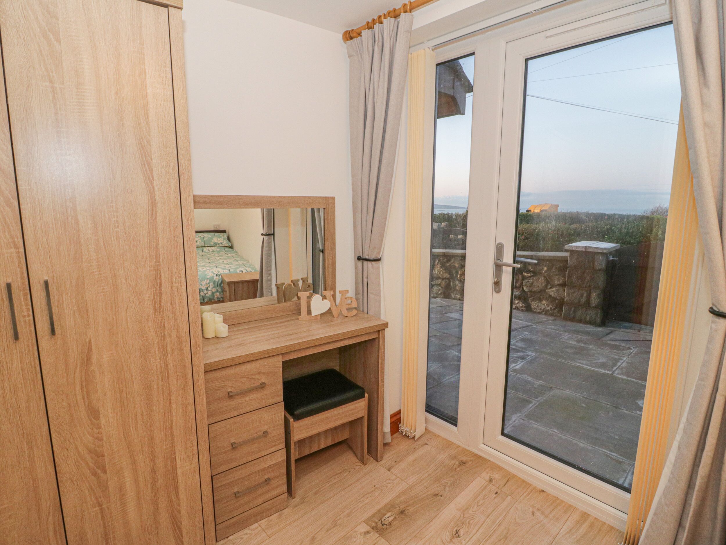 A bedroom with a wooden wardrobe, a desk with drawers, a mirror, and a sliding glass door leading to an outdoor area.