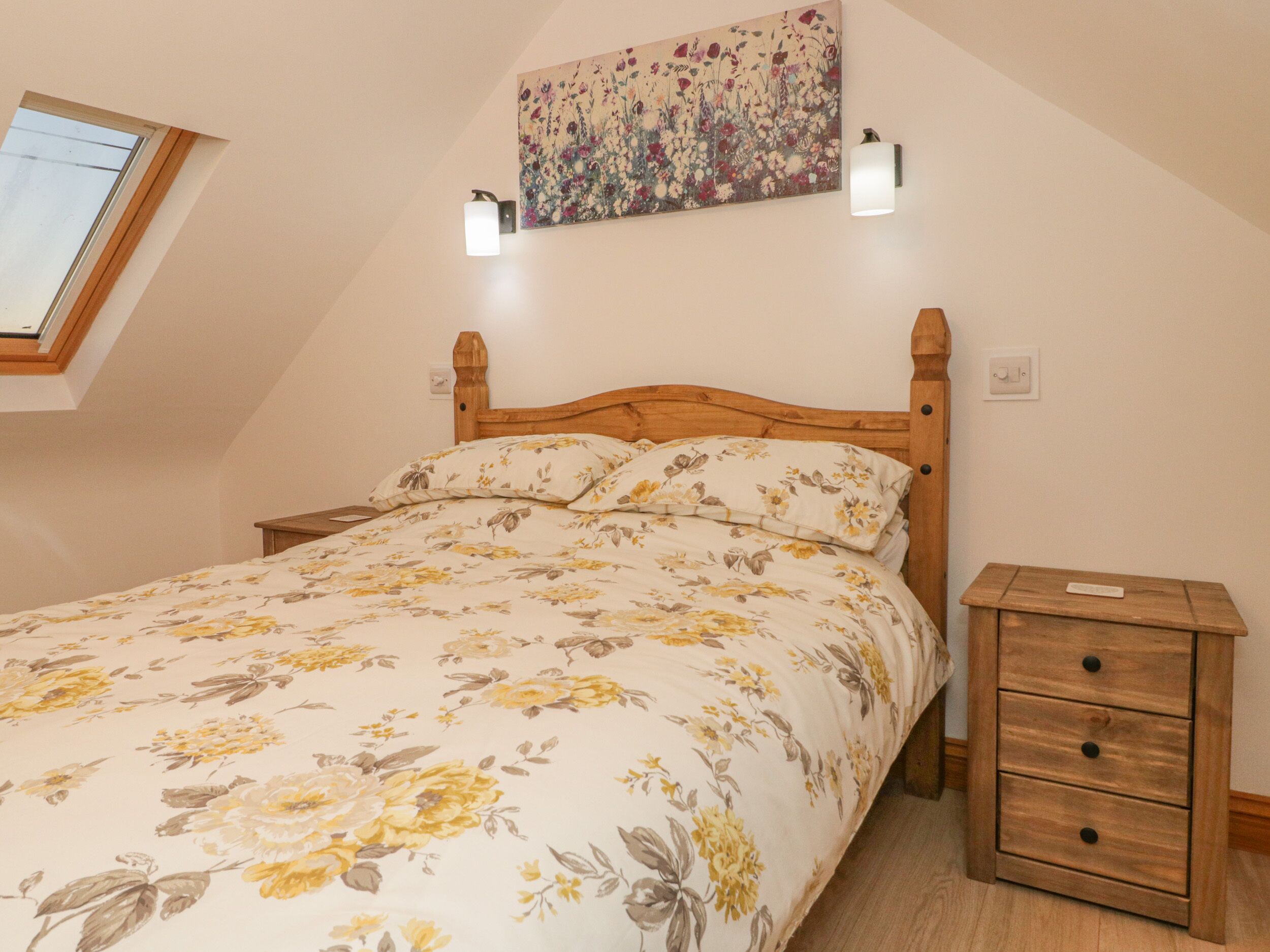 A bedroom with a sloped ceiling, a wooden bed frame, a floral bedspread, a bedside table with drawers, a wall-mounted light fixture, and a painting above the bed.