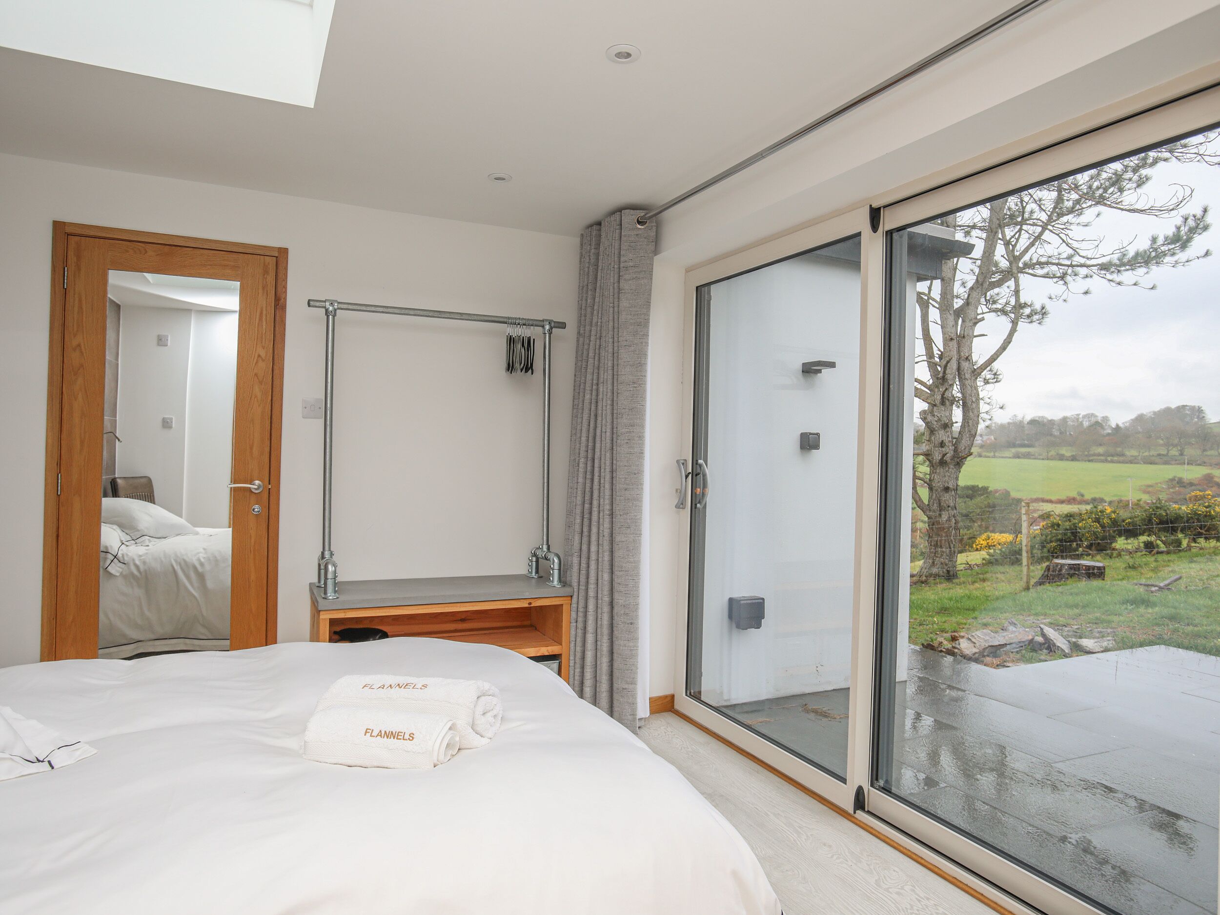 A modern bedroom with a large bed, a wooden bench, a glass door leading to a balcony, and a view of the countryside.
