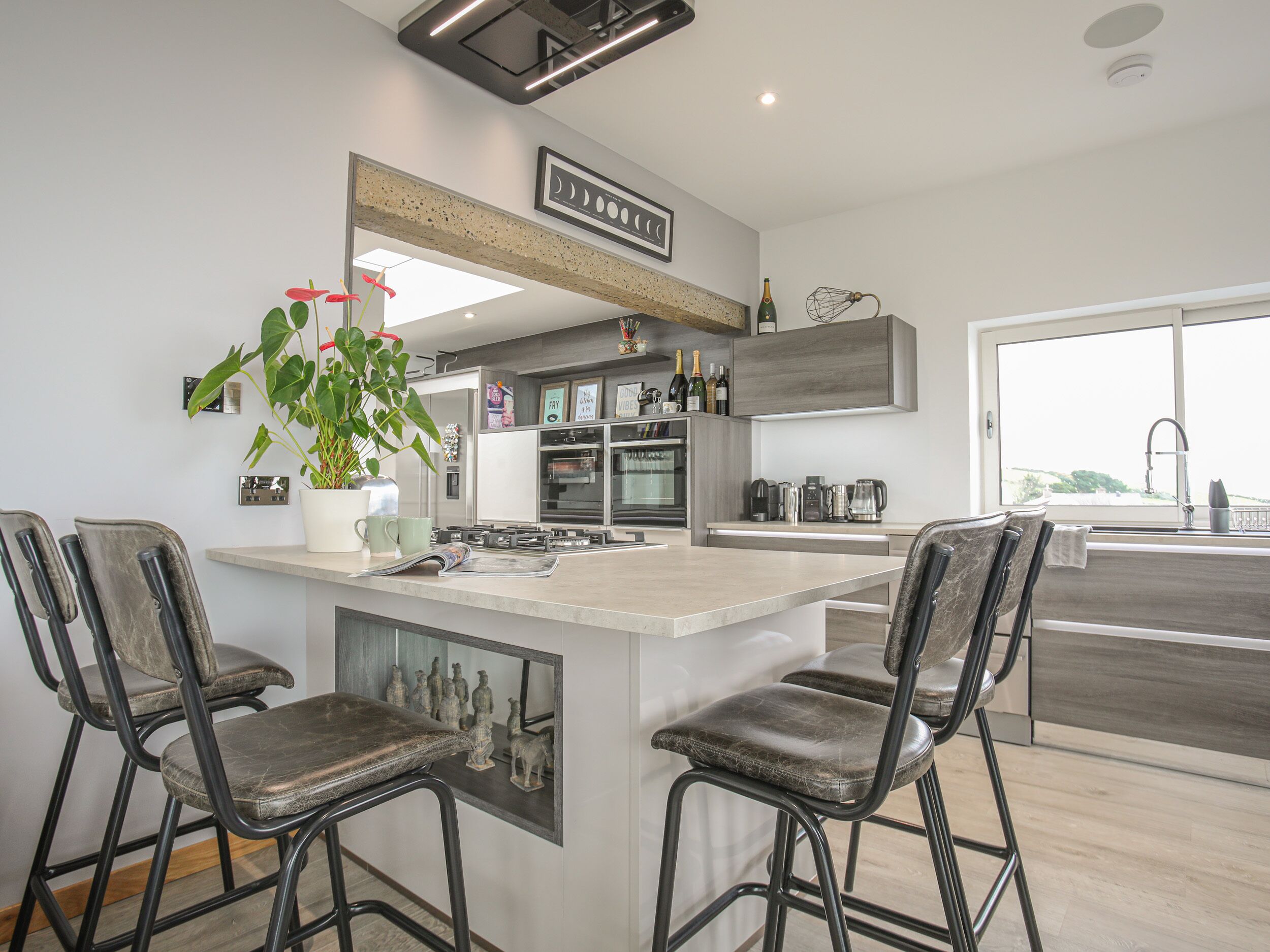 A modern kitchen with a central island, bar stools, a potted plant, and a view of the outdoors.