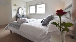 Une chambre à coucher avec un lit, une commode, un miroir, une fenêtre et un vase contenant une rose.
