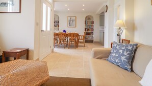 Un salon avec un canapé beige, un fauteuil en osier, un coin repas avec des chaises en bois, une bibliothèque et une lampe.
