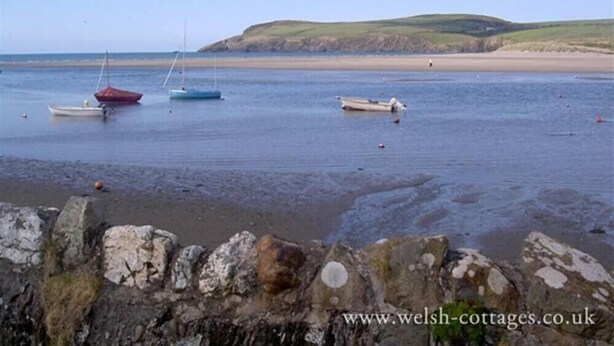 Un paysage côtier avec des bateaux sur une eau calme, une plage de sable et un mur de pierre au premier plan.