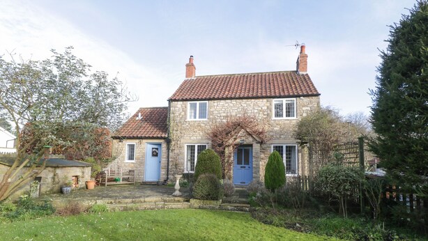 Une maison en pierre avec un toit recouvert de tuiles rouges, une porte bleue et un petit jardin avec une pelouse et quelques plantes en pot.