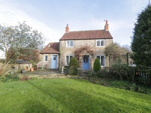 A stone house with a red-tiled roof, a blue door, and a small garden with a lawn and a few potted plants. - Howe Green Cottage (York)