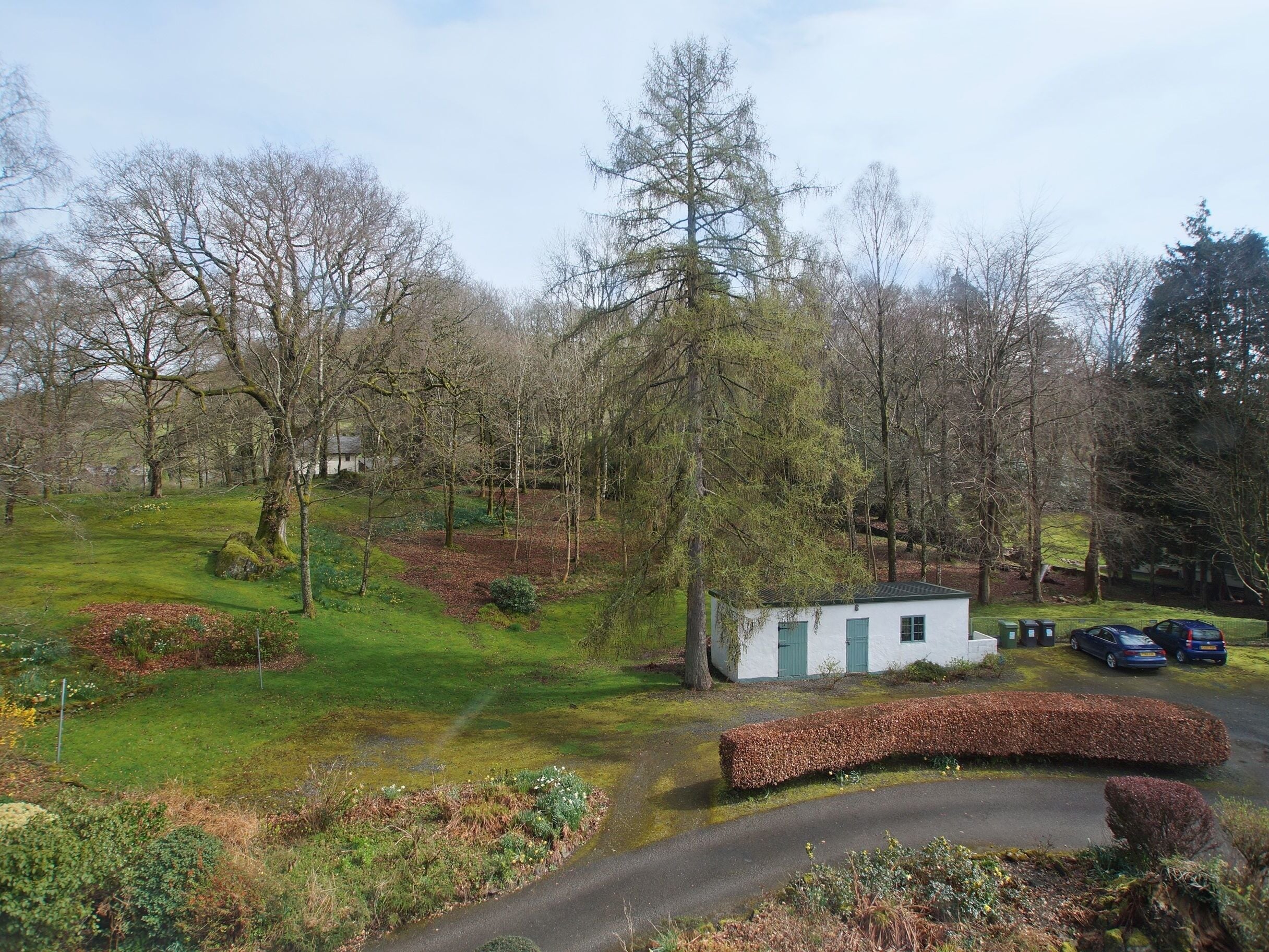 A small white building with green shutters, surrounded by a lush garden and trees.