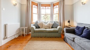 A cozy living room with a green sofa, a grey sofa, and a small table. There are curtains on the windows, and a radiator is visible on the wall. - Wisteria (St Austell)