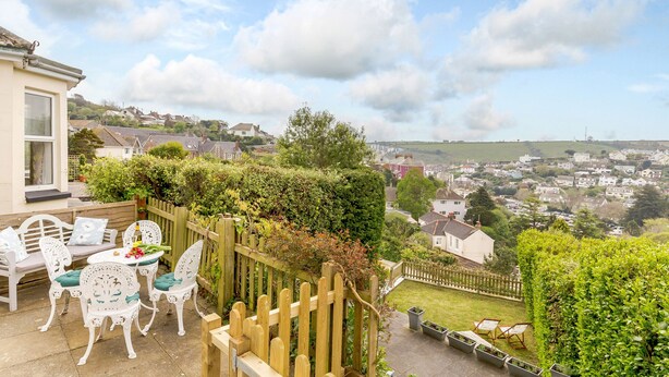 A patio with white chairs and a table, overlooking a residential area with houses and a hill in the distance. - Wisteria (St Austell)
