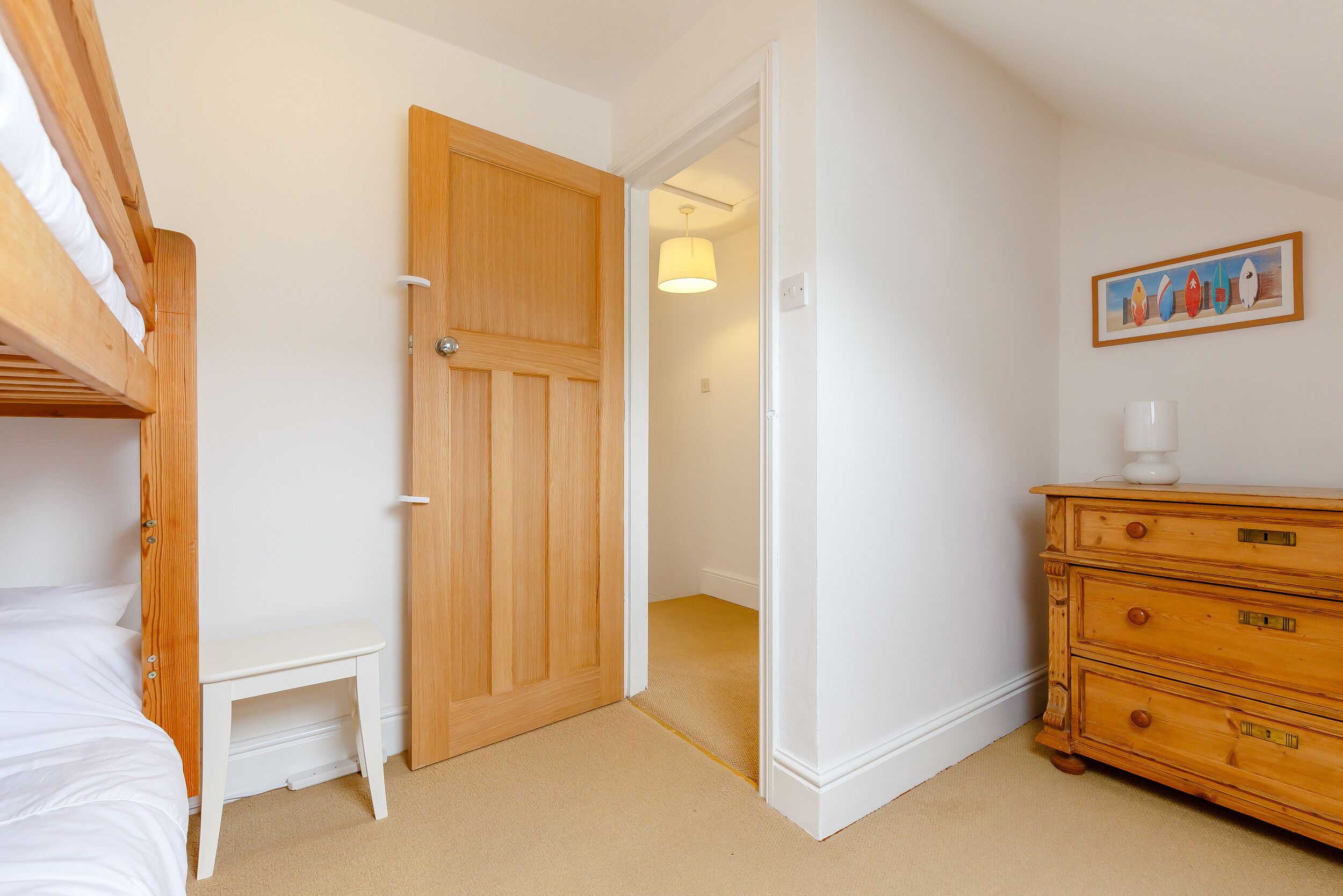 A bedroom with a bunk bed, a wooden dresser, a small bedside table, and a door leading to another room.