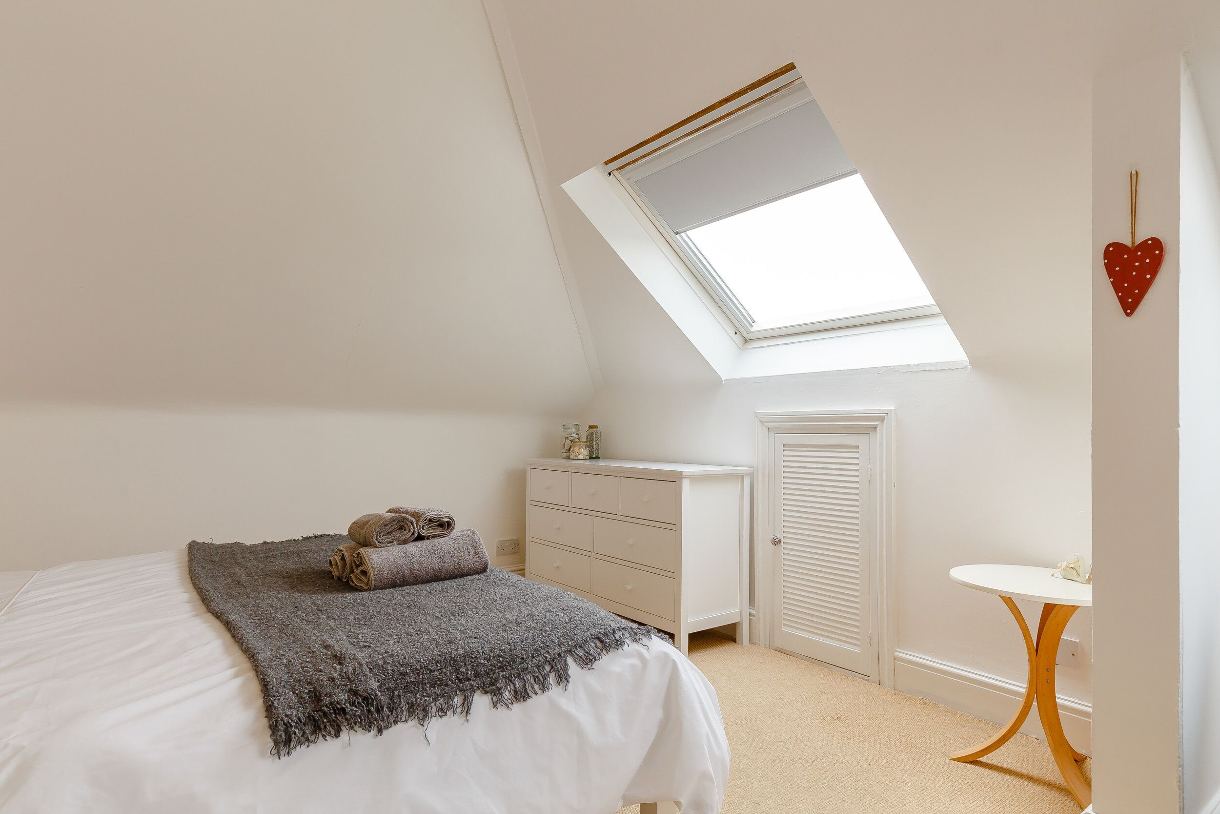 Une chambre avec un plafond en pente, une lucarne, un lit recouvert d’une couverture grise, une commode blanche et une petite table ronde.