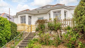 A two-story house with a stone staircase leading to the front door, surrounded by greenery and a wooden fence. - Wisteria (St Austell)