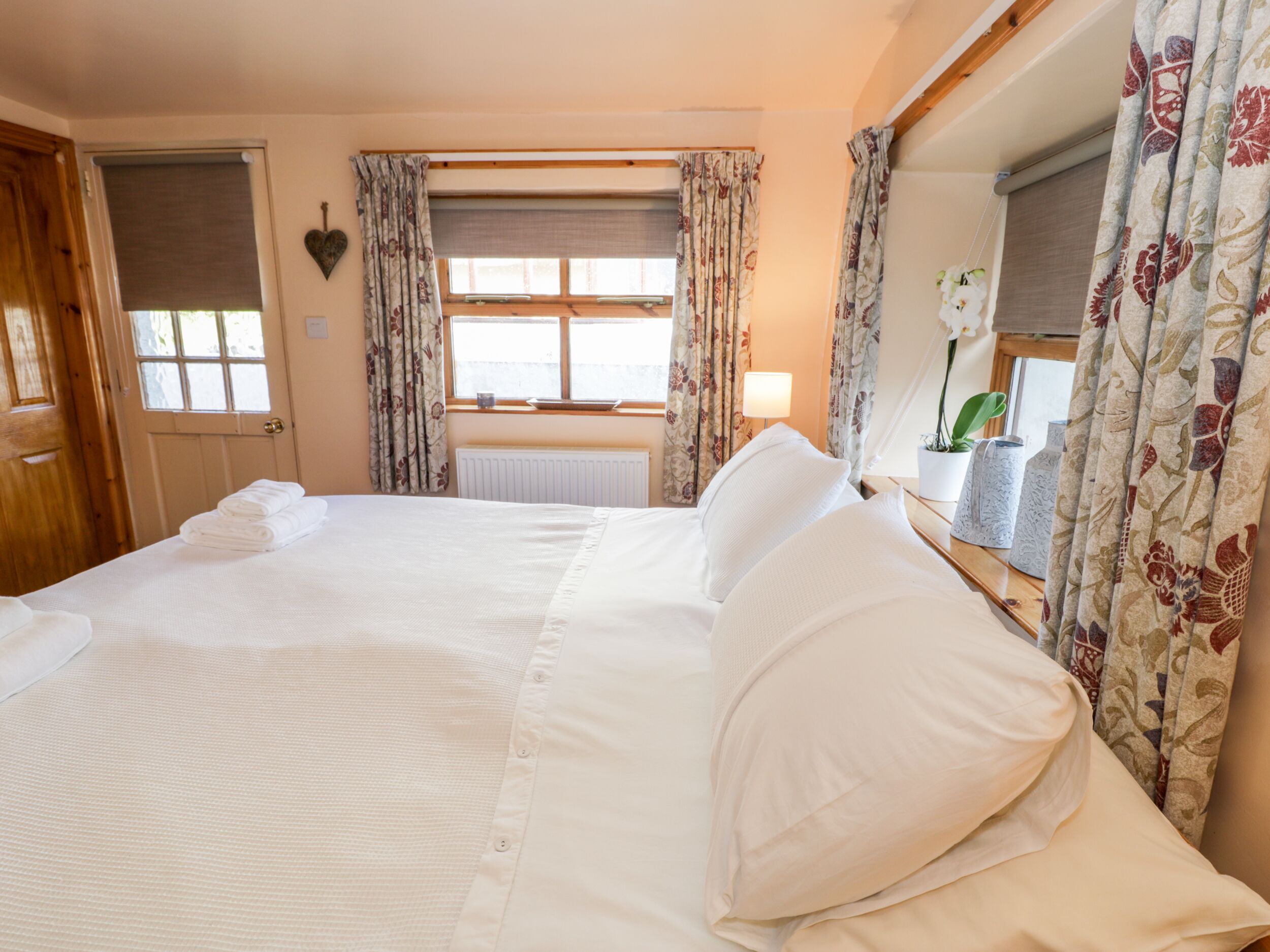 A large bed with white linens, a wooden door, and a window with floral curtains.