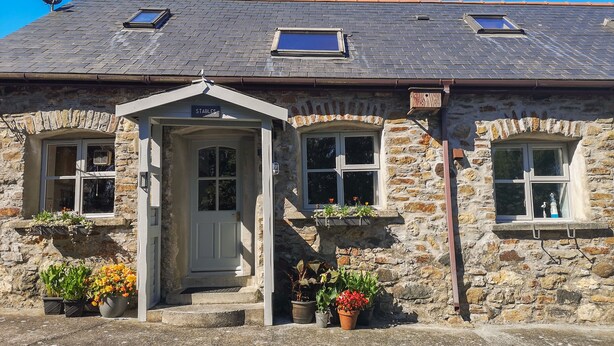Une maison en pierre avec une porte blanche, deux fenêtres ornées de jardinières et une antenne parabolique.
