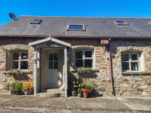 A stone house with a white door, two windows with flower boxes, and a satellite dish. - The Stables (Llanerchymedd)