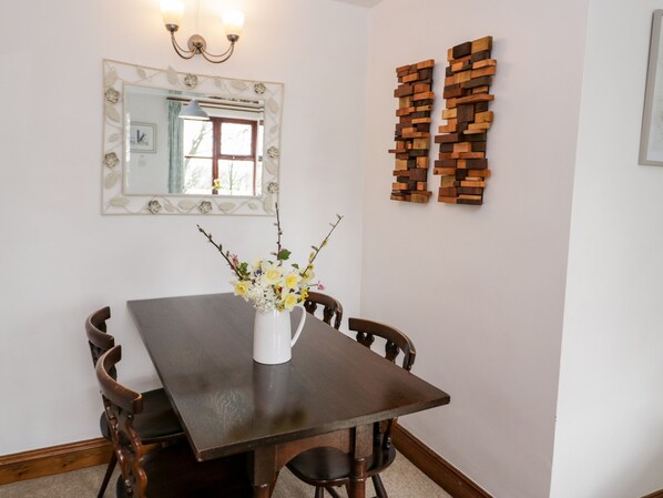 A dining area with a wooden table, chairs, a vase with flowers, a mirror, and wooden wall art. - The Stables (Llanerchymedd)