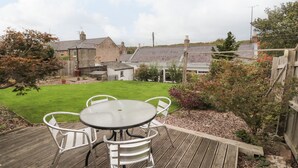A patio with a round table and four chairs, surrounded by a wooden deck and a grassy lawn. - Victoria Cottage (Berwick-upon-Tweed)