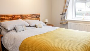 A neatly made bed with a wooden headboard, a bedside lamp, and a window with curtains. - Victoria Cottage (Berwick-upon-Tweed)