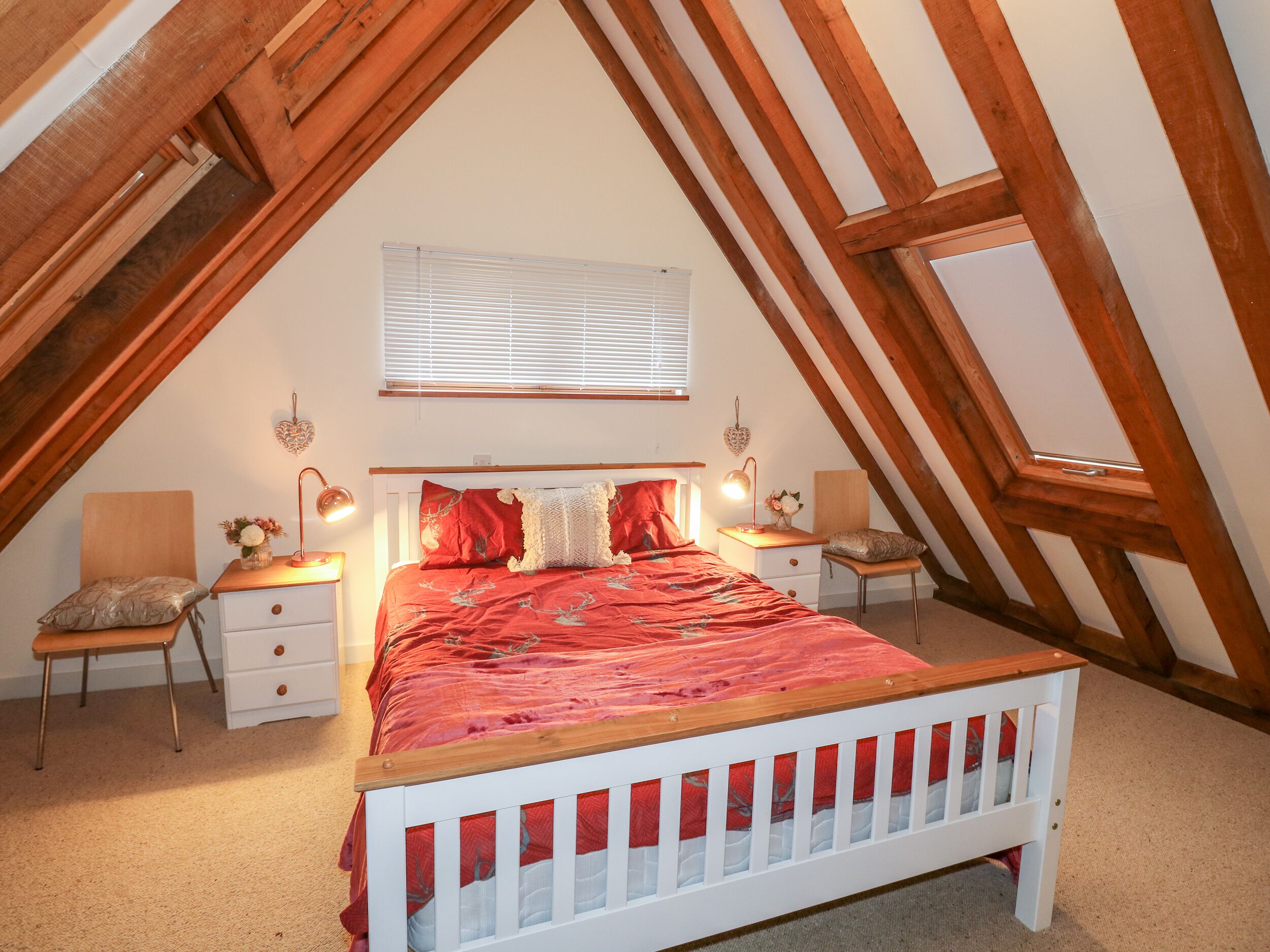 A bedroom with a sloped ceiling, a wooden bed frame, a red bedspread, two bedside tables with lamps, and a window with blinds.