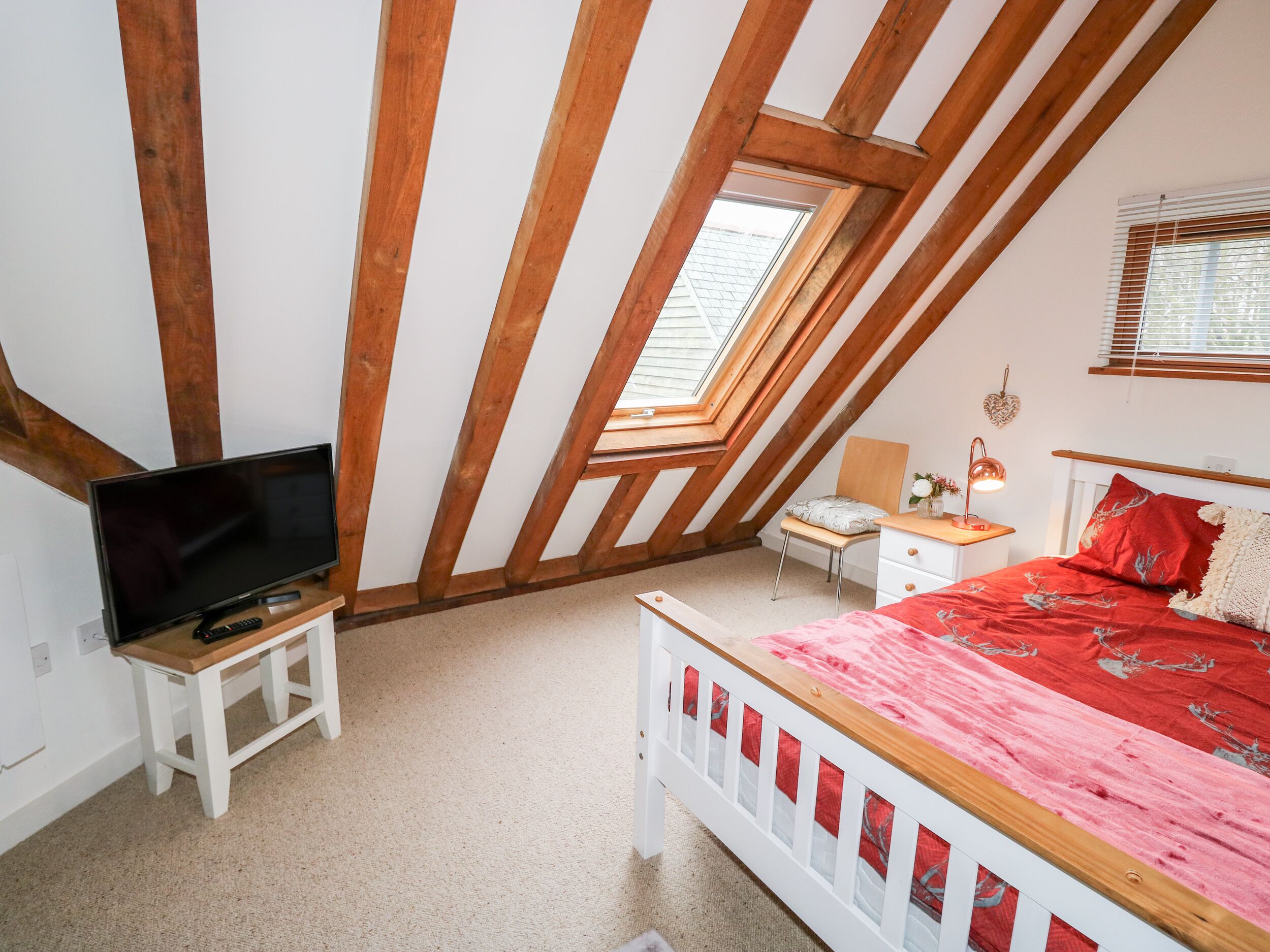A bedroom with a sloped ceiling, a bed with a red and white bedspread, a television on a small table, and a desk with a lamp.