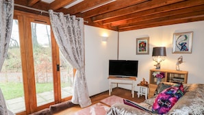 Ein gemütliches Wohnzimmer mit Fernseher auf einem weißen Schrank, einem Holzschrank mit Dekorationen und einem großen Fenster mit Vorhängen.