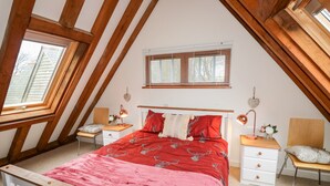 Ein Schlafzimmer mit Schrägen, einem Bett mit roter Decke, zwei Nachttischen mit Lampen und einem Fenster mit Jalousien.