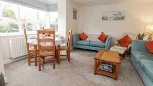 Ein Wohnzimmer mit Esstisch, zwei Holzstühlen, einem blauen Sofa mit orangefarbenen Kissen, einem Holztisch und einer Heizung.