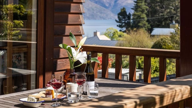 Um deck de madeira com uma mesa posta para refeição, uma garrafa de vinho e um vaso com flores.