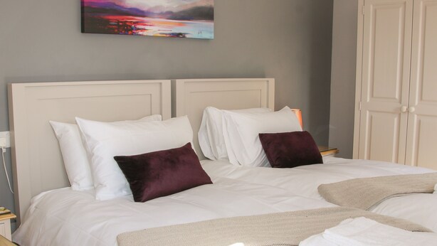 Una cama bien hecha con sábanas blancas y dos almohadas oscuras, una manta beige y una toalla blanca doblada al pie de la cama.