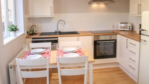 Una cocina moderna con armarios blancos, encimera de madera, horno empotrado y una zona de comedor para dos personas con sillas blancas y un mantel individual rosa.