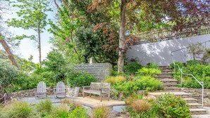 A garden patio with a wooden bench, a Adirondack chair, and a stone staircase leading up to a wooden deck. - 3 Rose Cottages (St. Agnes)