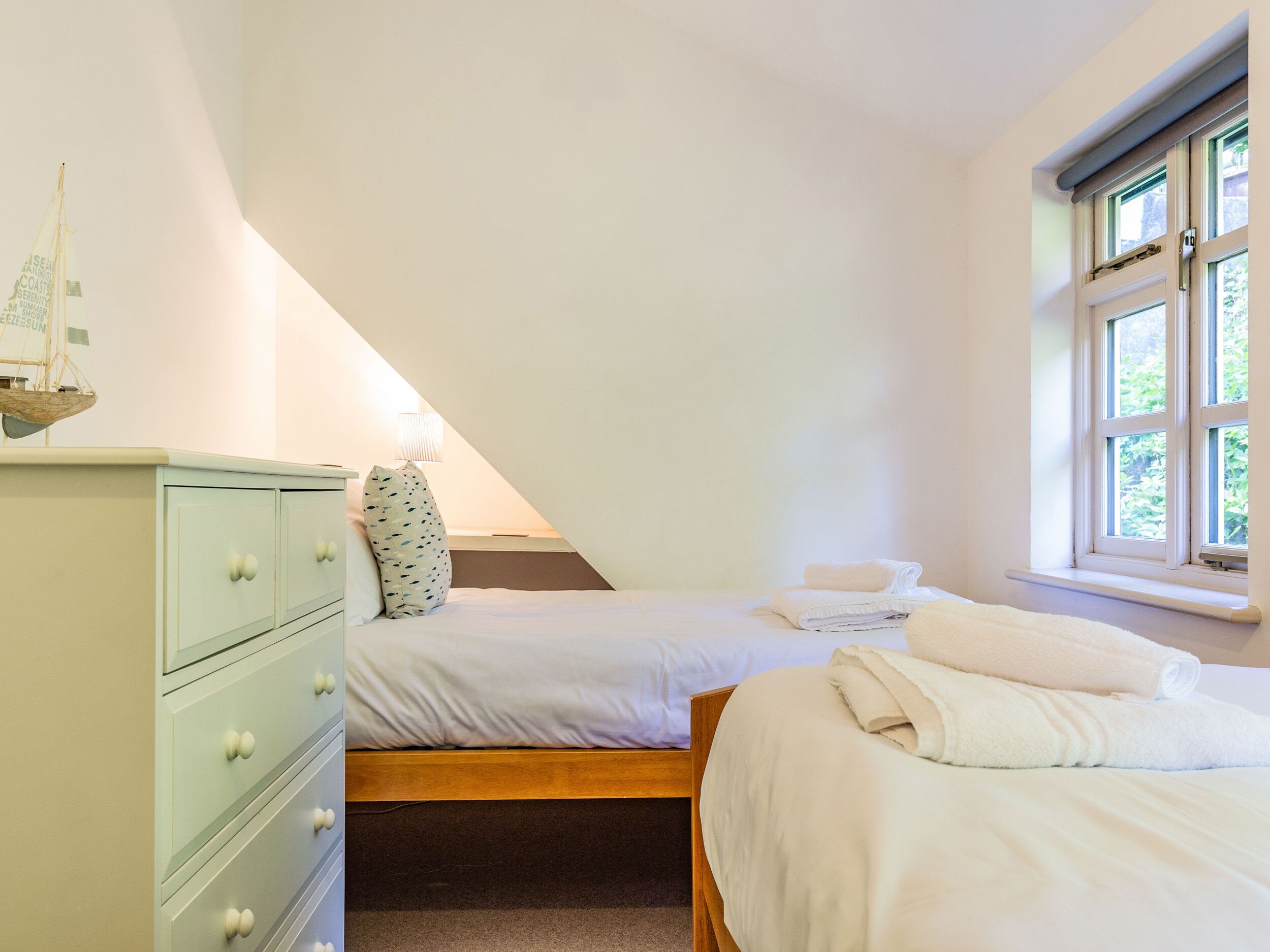 A bedroom with a sloped ceiling, a bed with white bedding, a nightstand, a dresser with drawers, and a window with a view of greenery.