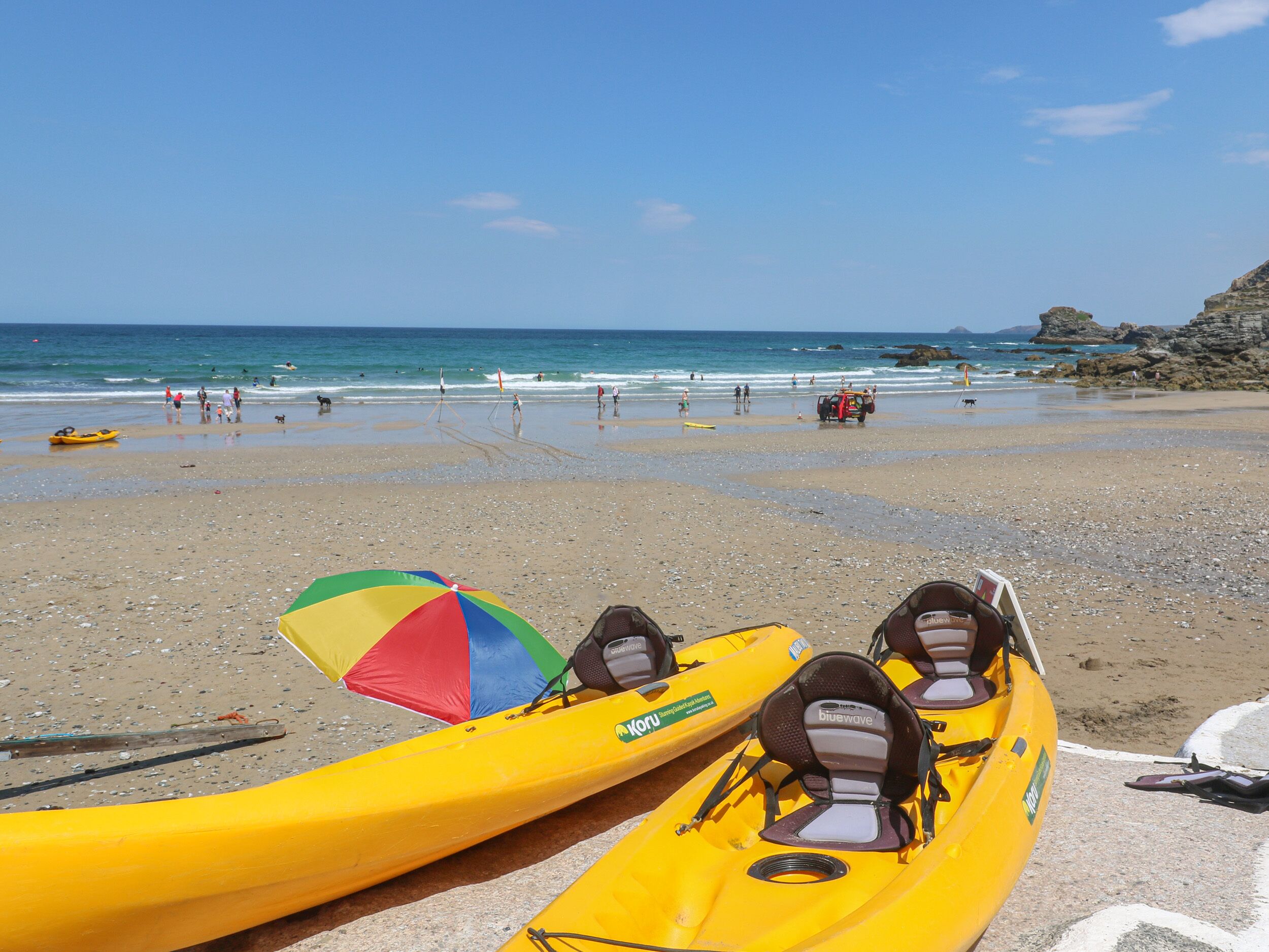 A beach with kayaks lined up on the sand, a colorful umbrella, and a rocky cliff in the background.