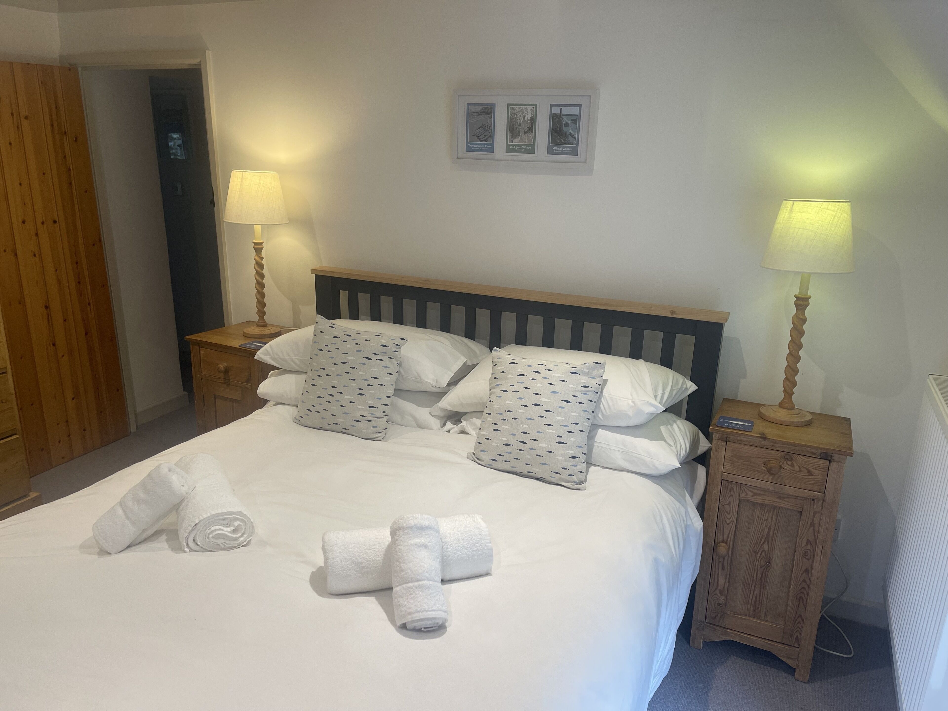 Una cama bien hecha con sábanas blancas y almohadones decorativos, dos mesitas de noche con lámparas y un cuadro enmarcado sobre la cama.