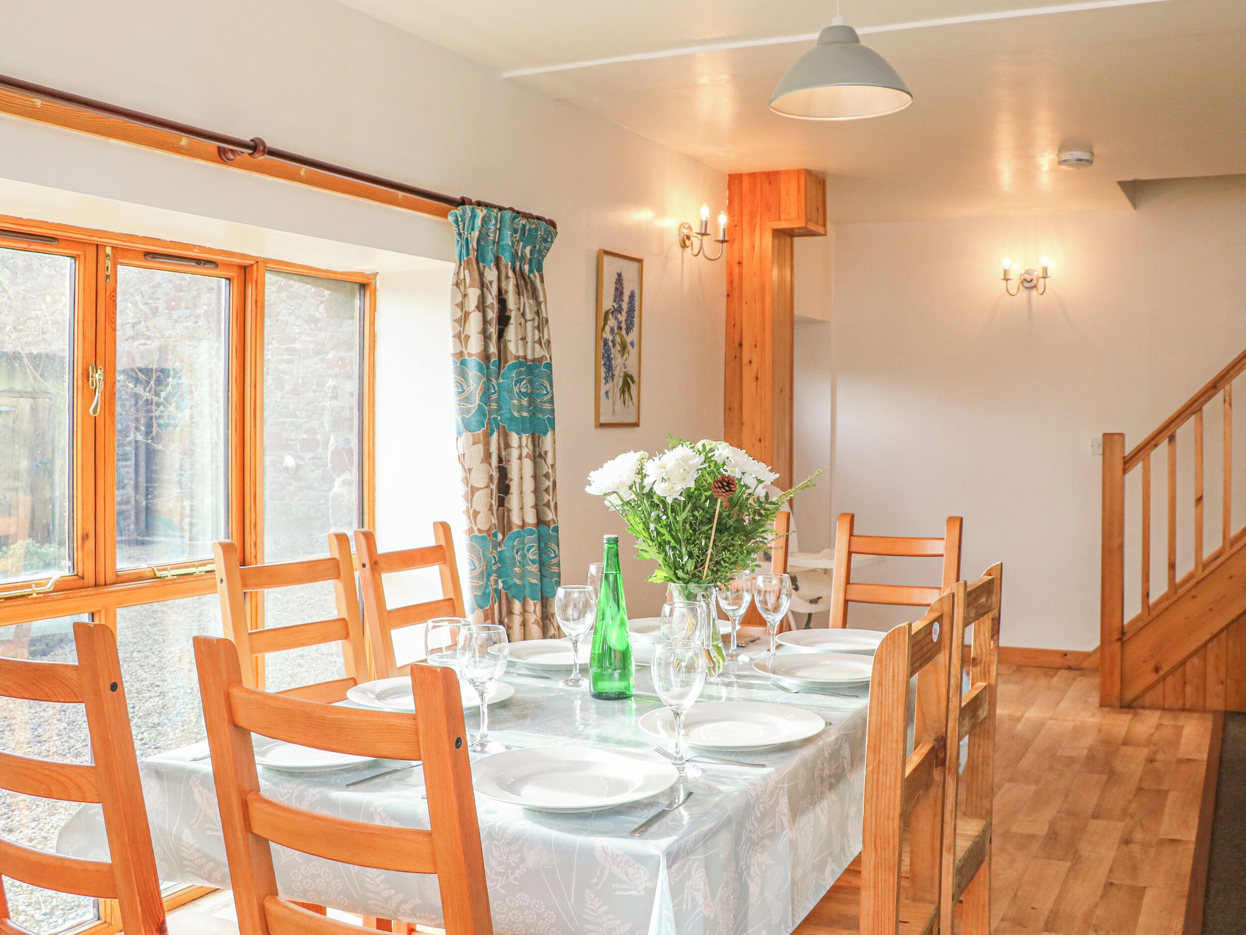 A dining area with a long table set for four, wooden chairs, a vase with flowers, and a green bottle.