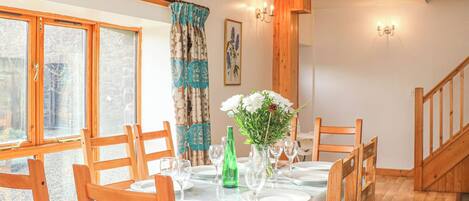Un coin repas avec une longue table dressée pour quatre personnes, des chaises en bois, un vase rempli de fleurs et une bouteille verte.