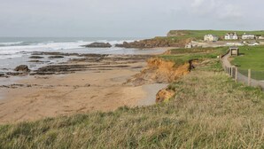 Un paysage côtier avec une plage de sable, des affleurements rocheux et des zones herbeuses. On y trouve des maisons et un sentier qui longe le bord de la falaise.