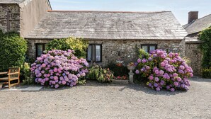Un bâtiment en pierre avec une allée de gravier et de grands buissons d’hortensias violets devant.