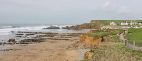 A coastal landscape with a sandy beach, rocky outcrops, and grassy areas. There are houses and a pathway along the cliff edge.