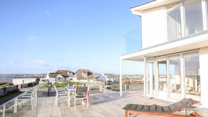 Una terrazza esterna moderna con una poltrona da relax, un tavolo con sedie e una ringhiera in vetro che si affaccia su una zona costiera con case sullo sfondo.