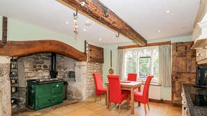 A rustic kitchen with a stone fireplace, wooden beams, and a dining table with red chairs. - Chapel House (Stonehouse)