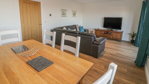 A modern living room with a wooden dining table, white chairs, a dark leather sofa, a TV on a wooden cabinet, and a wooden door. - Isallt (Caernarfon)