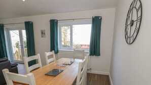 A dining area with a wooden table, white chairs, and a view of the outdoors through a window with teal curtains. - Isallt (Caernarfon)