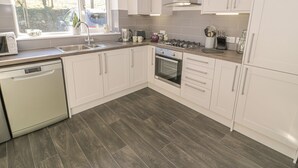 A modern kitchen with white cabinets, a stainless steel range hood, a dishwasher, and a window with blinds. - Isallt (Caernarfon)