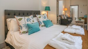 A neatly made bed with patterned pillows, a bedside table with a lamp, and a view into a living area with a sofa and a table. - Lakeside Cottage (Macclesfield)