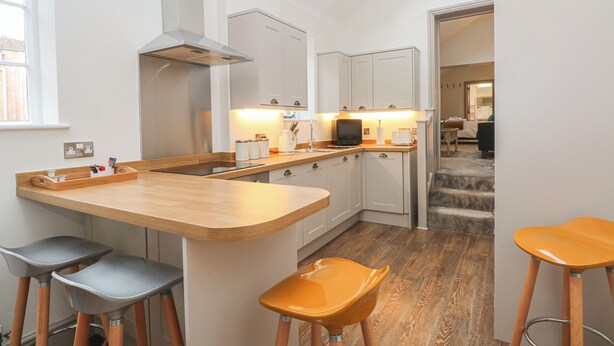 A modern kitchen with a central island, bar stools, and a view of the dining area. - Lakeside Cottage (Macclesfield)